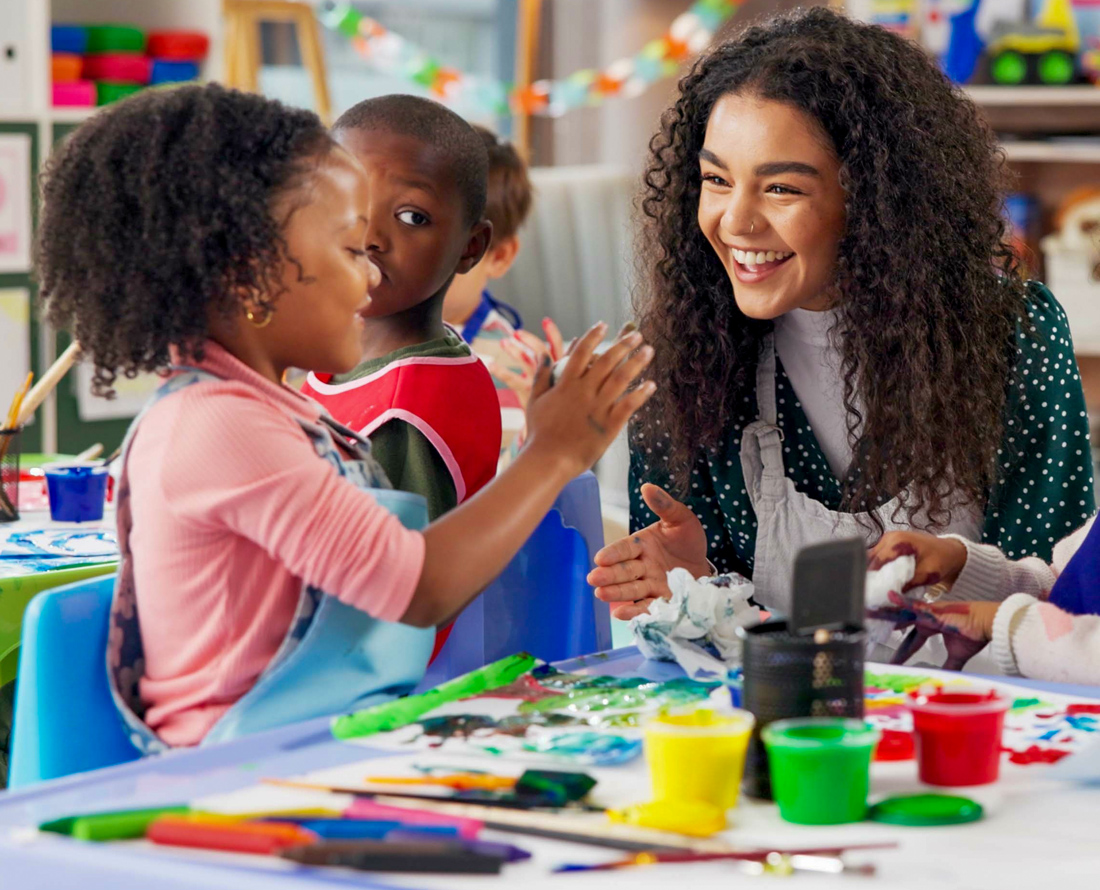 Teacher engages with primary school pupil in class. Art resources including paints sit on the table.
